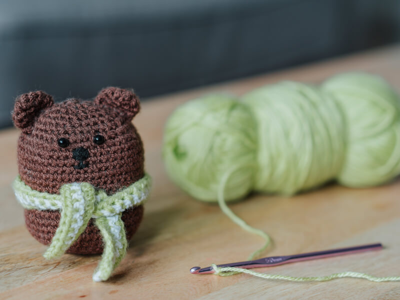 A crocheted teddy sat next a crochet hook and yarn